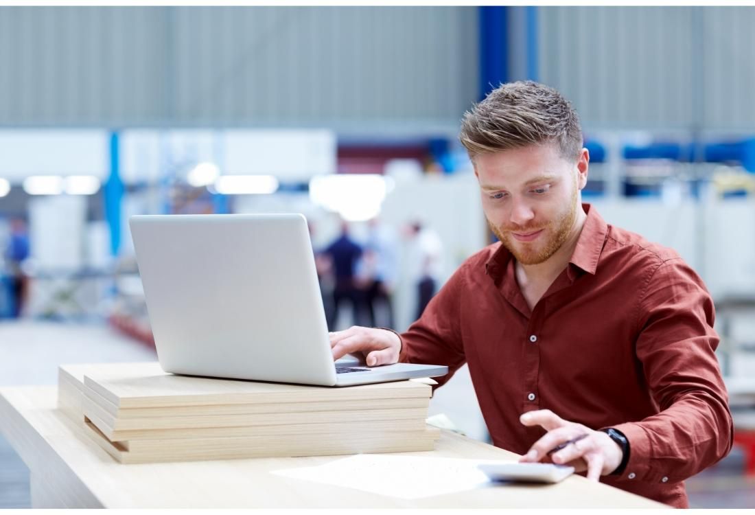 Young man in a red shirt sat at a desk in a warehouse keeping track of one of his customers' stock on a laptop whilst using a calculator.